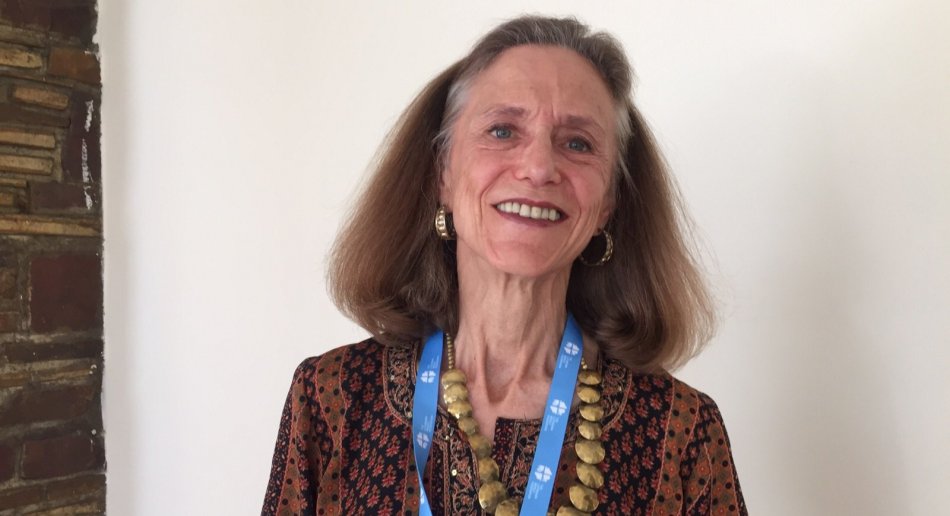 A white woman with graying hair stands against a white wall, smiling.